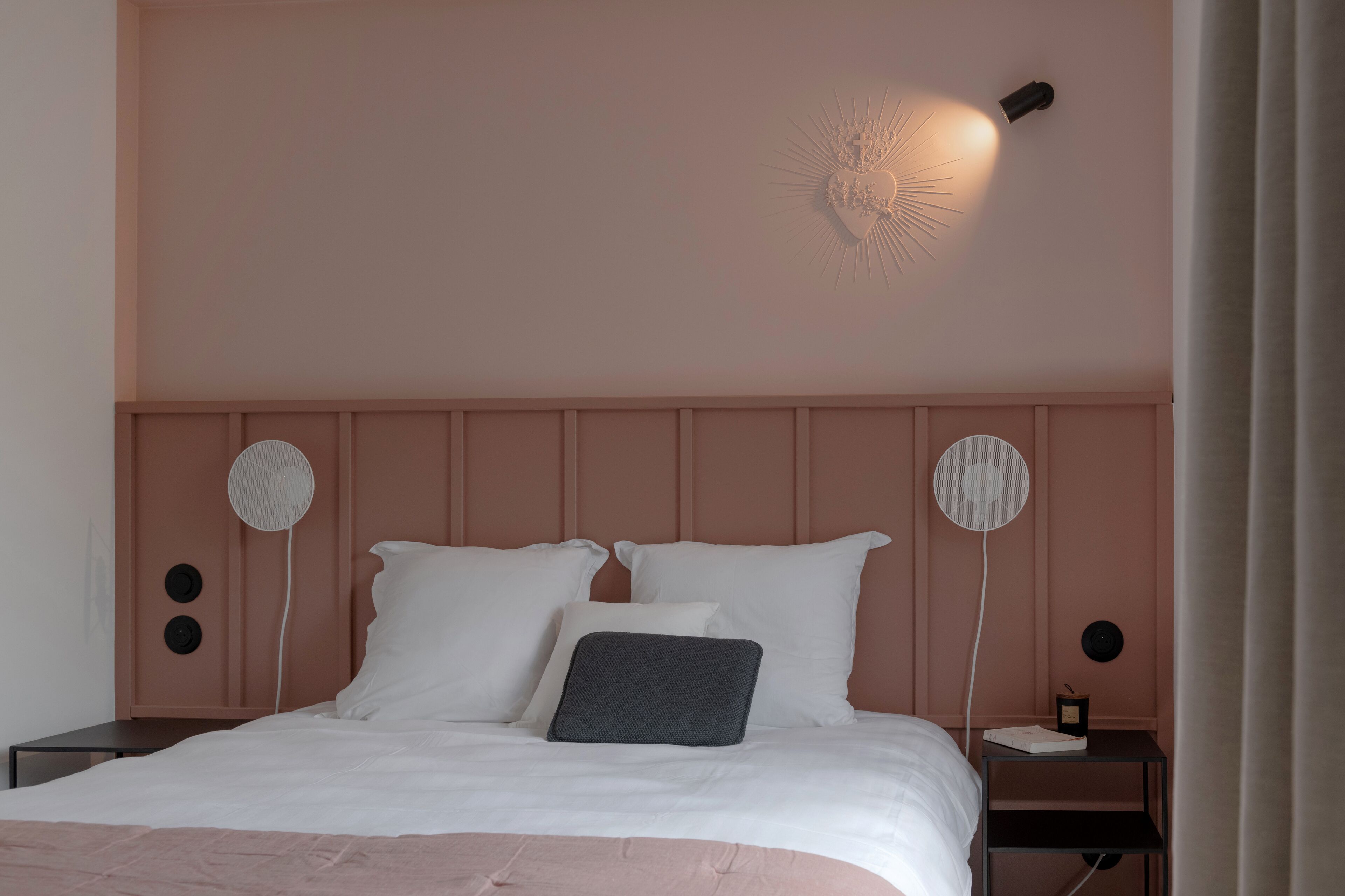Una cama bien hecha con sábanas blancas y un almohadón gris, dos mesitas de noche con lámparas blancas, y una lámpara de pared.