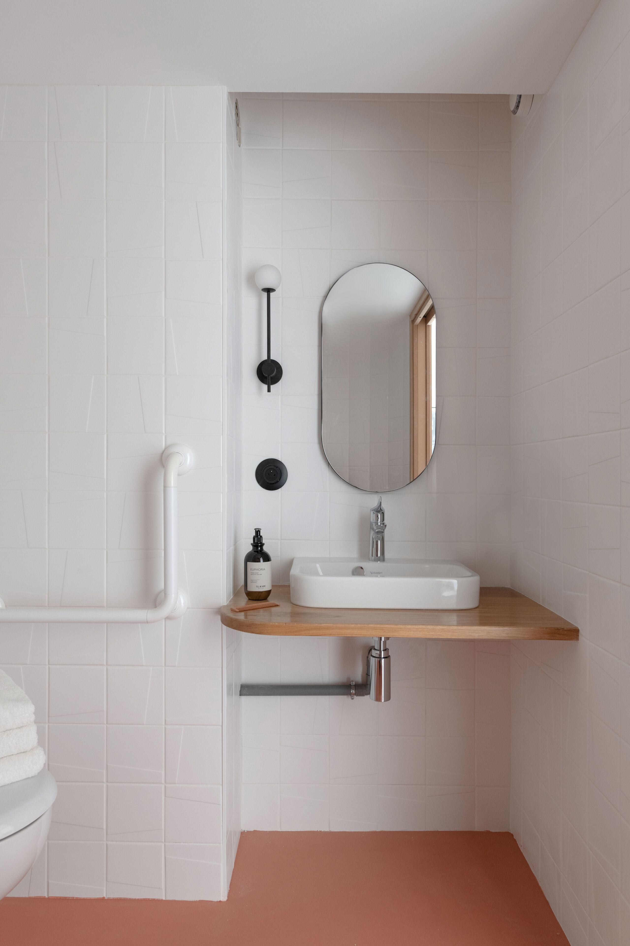 Een badkamer met witte betegelde wanden, een houten wastafelmeubel, een witte wasbak, een spiegel en een toilet.