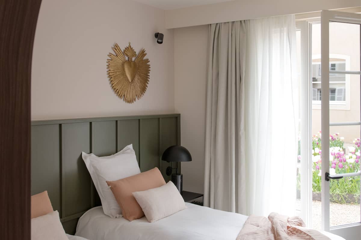 A hotel room with two beds, a green headboard, a wall-mounted heart-shaped decoration, a nightstand with a lamp, and a view of a balcony with flowers.