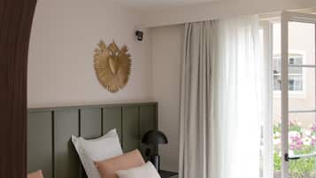 A hotel room with two beds, a green headboard, a wall-mounted heart-shaped decoration, a nightstand with a lamp, and a view of a balcony with flowers.