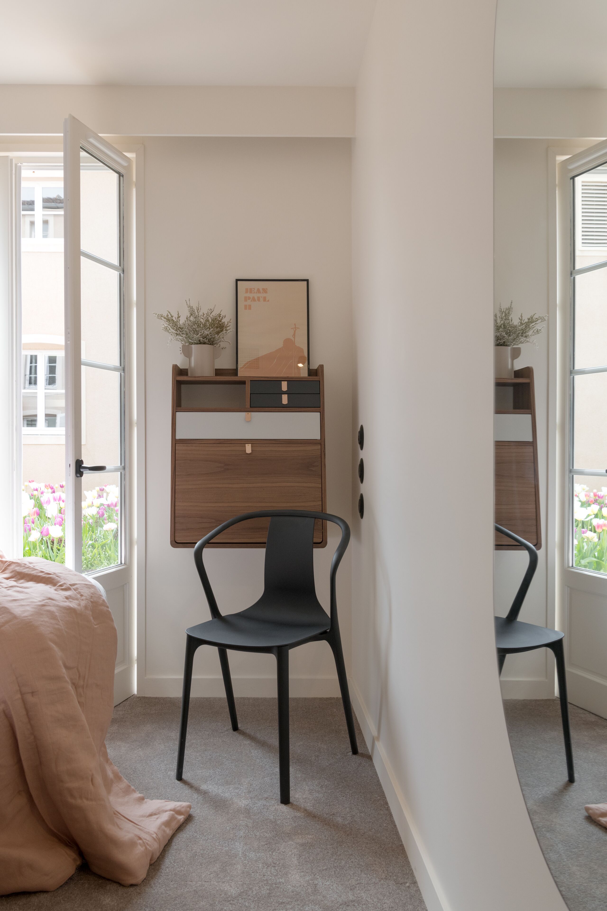 Een slaapkamer met een bed, een bureau met stoel, een raam met uitzicht op bloemen en een deur naar buiten.
