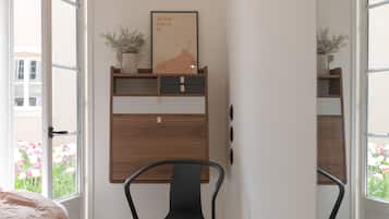 Een slaapkamer met een bed, een bureau met stoel, een raam met uitzicht op bloemen en een deur naar buiten.