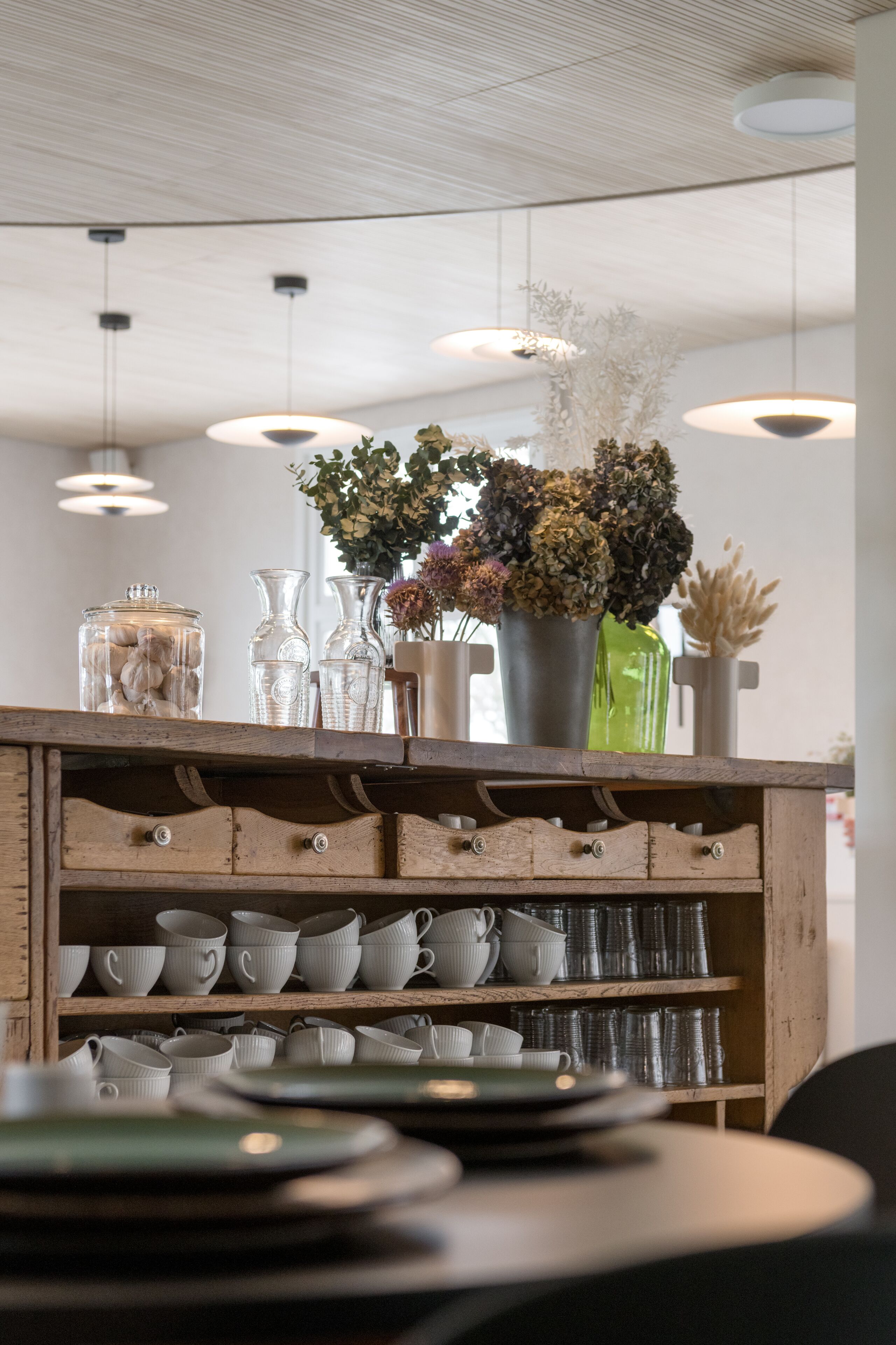Una estantería de madera con varios objetos de vidrio y cerámica, entre ellos tazas y platos, además de un jarrón con flores secas.