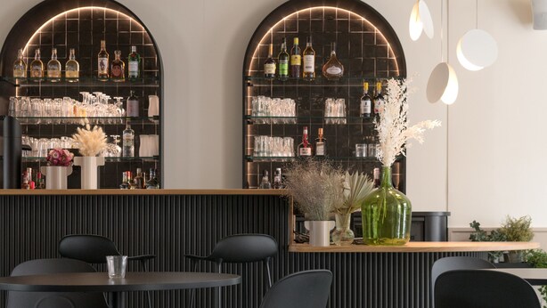 Un área de bar moderna con sillas negras, una barra de madera y estantes con botellas y vasos.