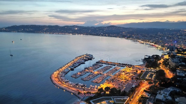 Vista aérea de un puerto deportivo con barcos amarrados, un estacionamiento y edificios a lo largo del frente marítimo.