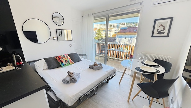 A modern hotel room with a bed, a small table with chairs, a balcony with a view of buildings, and a radiator.