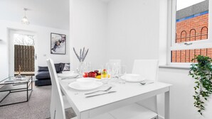 A modern dining area with a white table set for four, a glass coffee table, and a view of a brick building through a window. - The Marsons Collection - 2 Bed Apartment- Parking (Birmingham)
