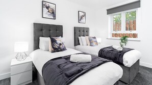 A modern hotel room with two beds, a nightstand, a window with a view of a wooden fence, and abstract artwork on the walls. - The Marsons Collection - 2 Bed Apartment- Parking (Birmingham)