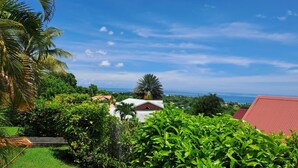 A lush garden with a view of the ocean, a red-roofed building, and a white building with a red roof. - Baobab 3 Vacation Rental – Tropical Charm & Ocean-View Pool (Sainte-Anne)