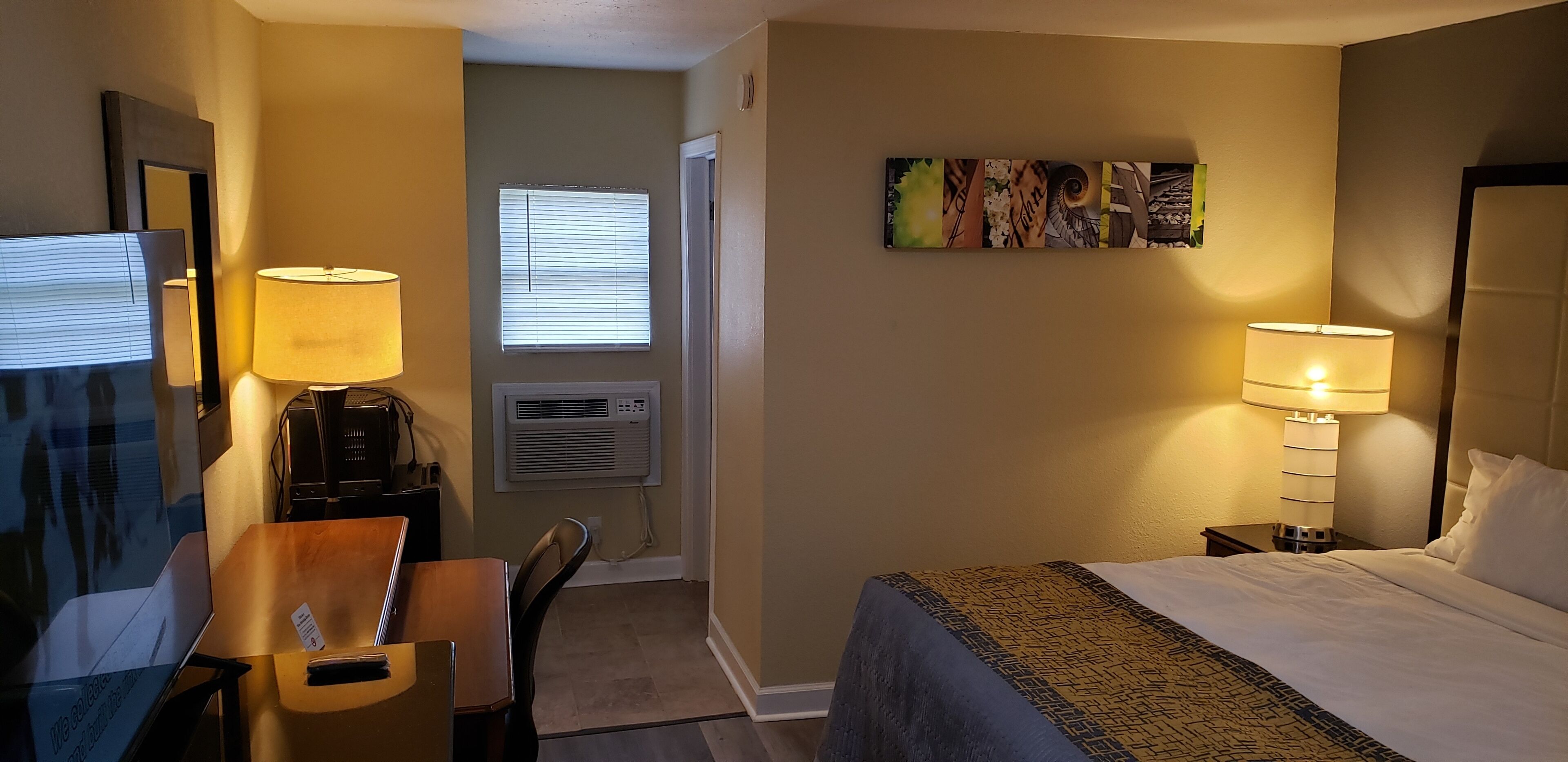 A hotel room with a bed, two bedside lamps, a desk with a chair, a TV, and a wall-mounted air conditioner.