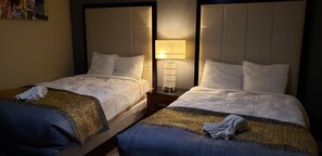 Two twin beds with white linens and a patterned blanket, a bedside table with a lamp, and a wall-mounted artwork.