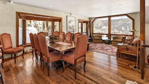 Un comedor espacioso con una mesa de madera y sillas a juego, ventanas amplias que ofrecen vistas a un paisaje nevado, y una zona de estar acogedora con un tapete estampado y un sofá.