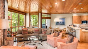A spacious living room with a large sectional sofa, two armchairs, and a glass coffee table. The room features wooden ceiling beams, large windows offering a view of greenery outside, and a kitchen area with wooden cabinets and a microwave.