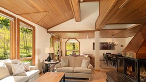 A cozy living room with a fireplace, wooden ceiling, and large windows offering a view of the outdoors.