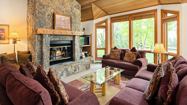 A cozy living room with a stone fireplace, wooden ceiling, and large windows offering a view of trees. - Sweeping down-valley views and comfortable upgrades. (Snowmass Village)