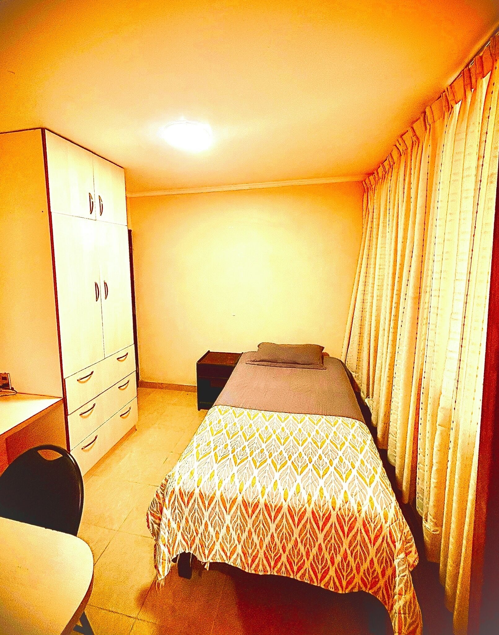Habitación pequeña con cama individual, escritorio y armario, con una colcha estampada y cortinas.