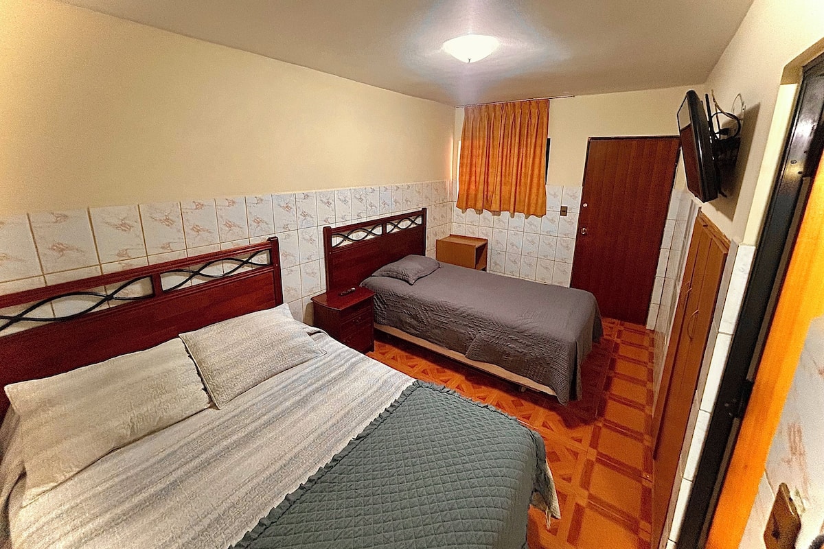 A small, compact room with two single beds, a wooden headboard, a small bedside table, a television mounted on the wall, and a door leading to another room.