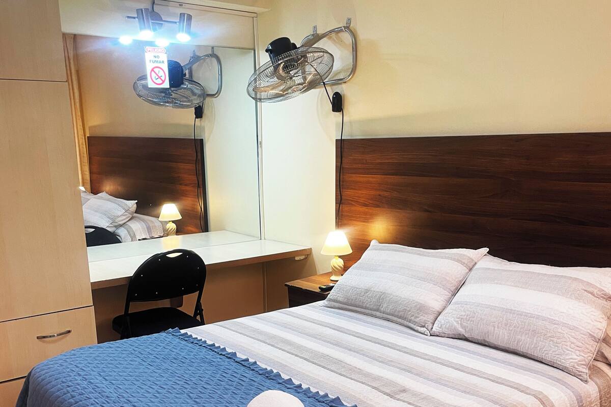A hotel room with a bed, a desk, a chair, a mirror, and a wall-mounted fan.