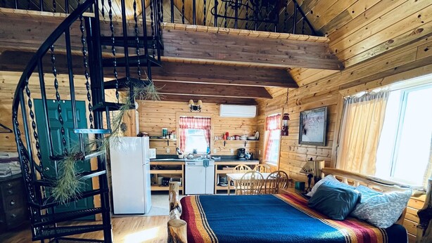 L’intérieur chaleureux d’un chalet avec un escalier en bois, un lit recouvert d’une couverture colorée, un coin repas et une cuisine.