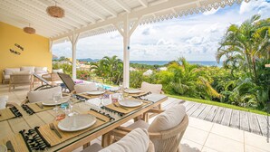 Uma área de refeições coberta ao ar livre, com uma mesa posta para quatro pessoas, com vista para uma paisagem tropical com palmeiras e o oceano.