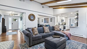A spacious living room with a grey sectional sofa, a matching ottoman, and a patterned rug. A dining area with a table and chairs is visible in the background.