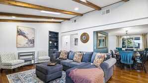 A spacious living room with a sectional sofa, a coffee table, and a patterned rug. There is a dining area with a table and chairs, and a large window with curtains.