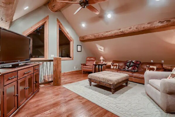 A cozy loft-style living room with a large flat-screen TV, leather furniture, and a wooden ceiling beam.