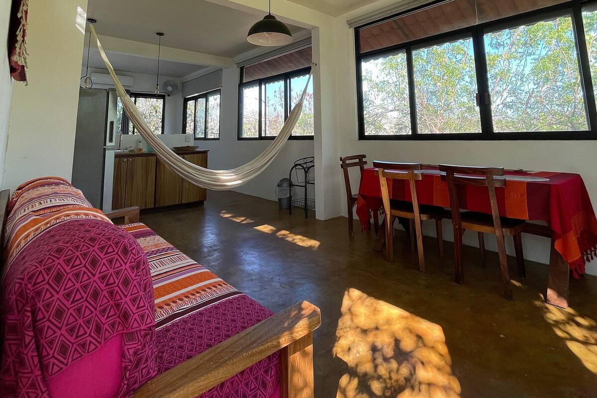 Un vaste espace de vie comprenant un hamac, une table à manger avec des chaises et de grandes fenêtres.