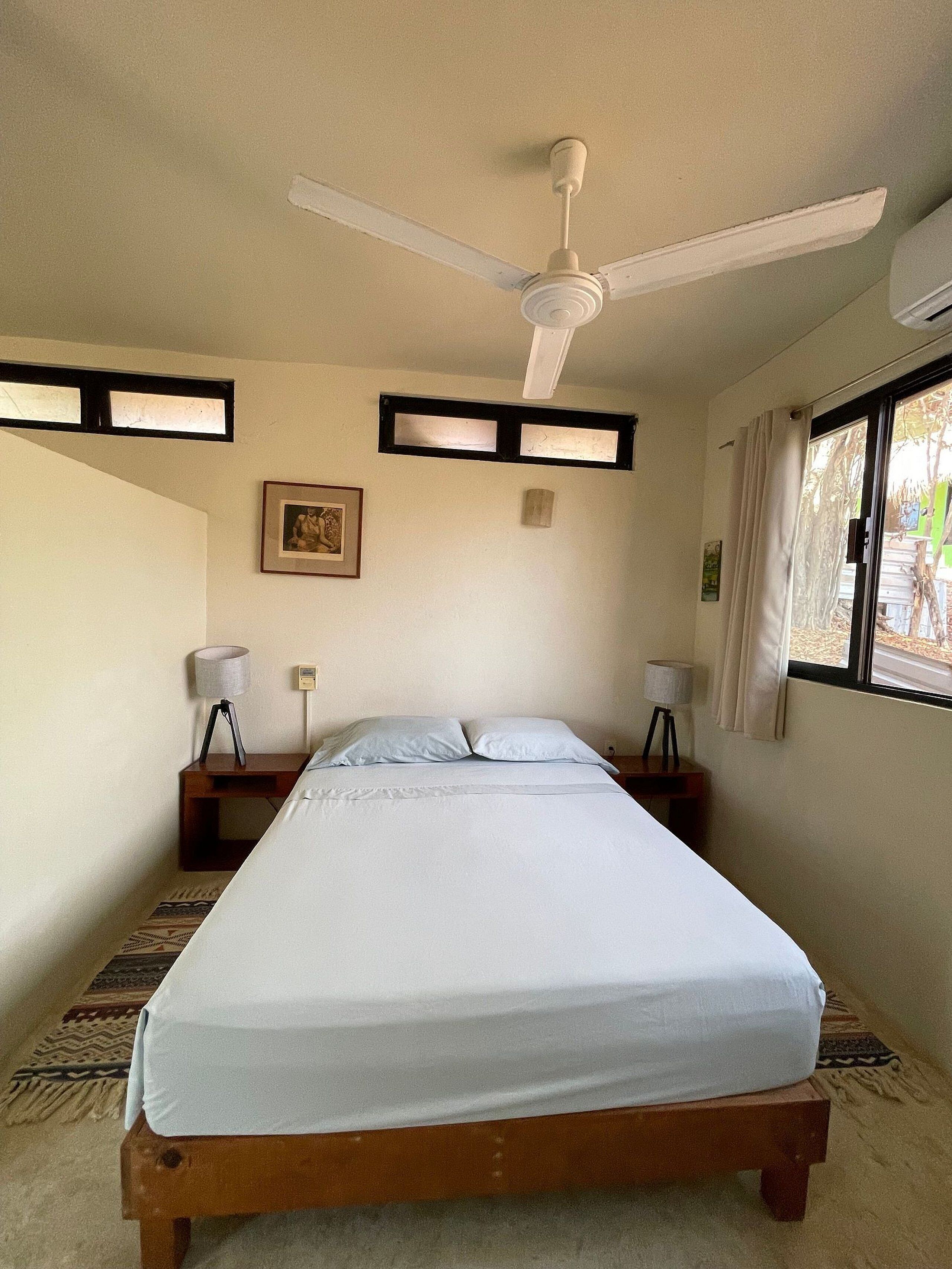 A bedroom with a single bed, a ceiling fan, two bedside tables with lamps, a framed picture on the wall, and a window with curtains.