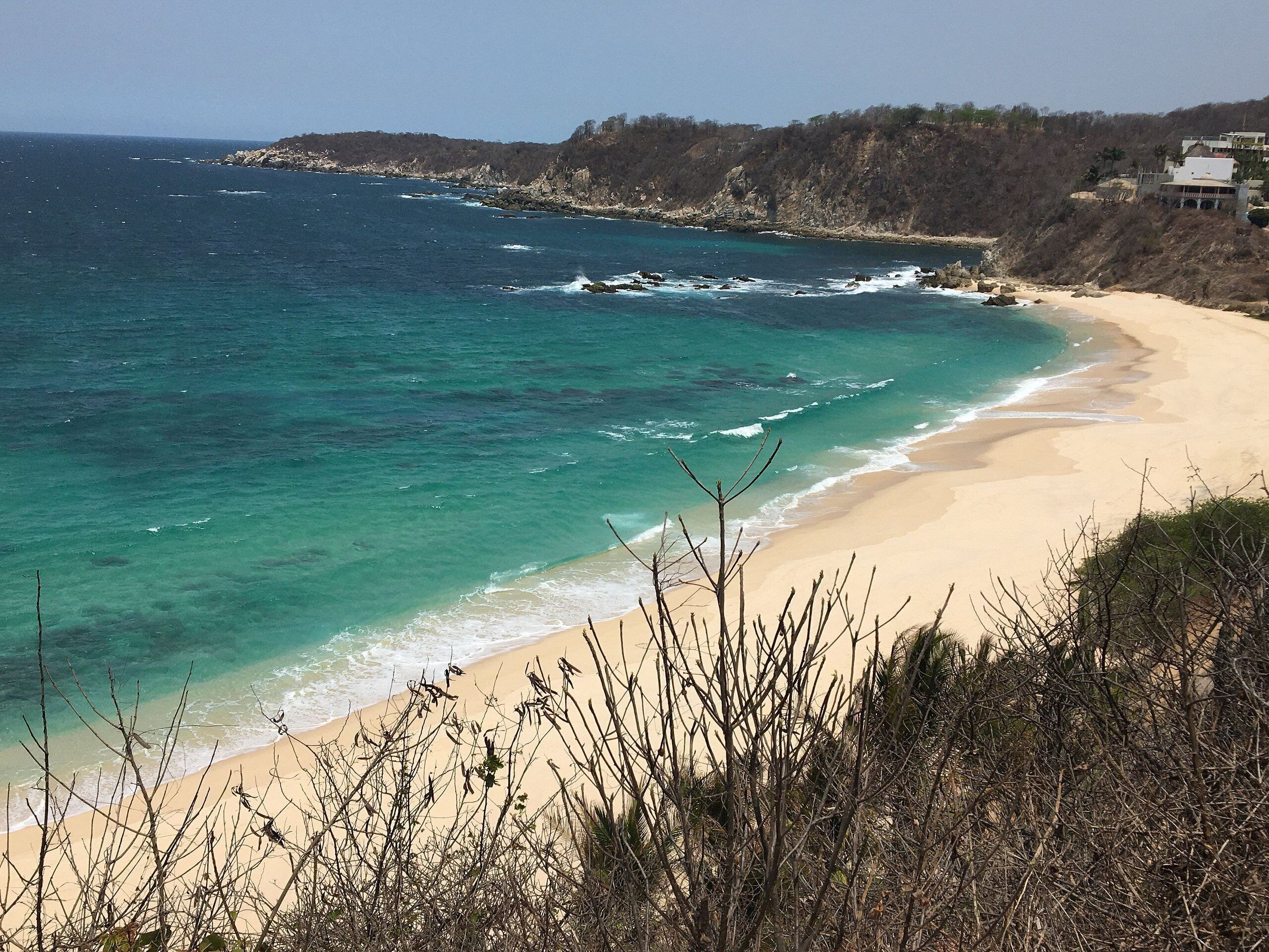 Uma praia com águas turquesa cristalinas, areal arenoso e falésias rochosas ao fundo.