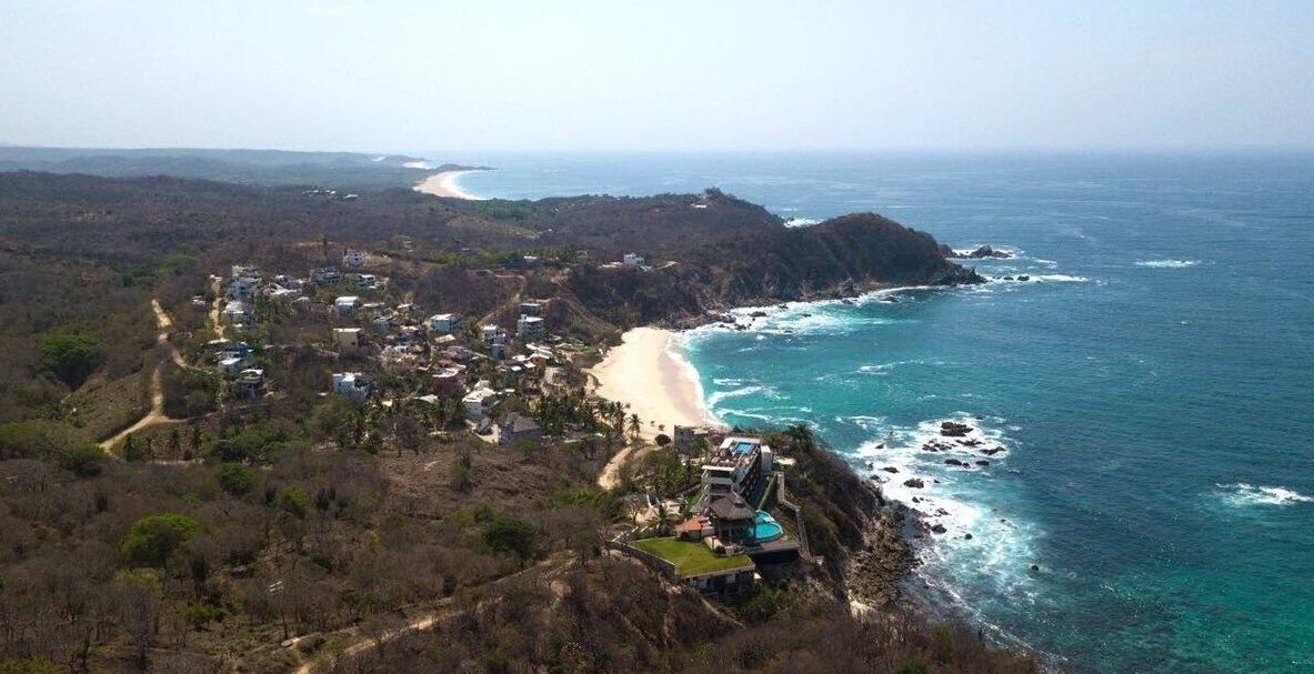 An aerial view of a coastal area with a beach, a cliffside with buildings, and a clear view of the ocean.