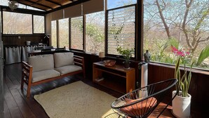 Un vaste espace de vie doté d’un plancher en bois, d’un canapé, d’un fauteuil et d’une table à manger. De grandes fenêtres offrent une vue sur les arbres et les plantes à l’extérieur.