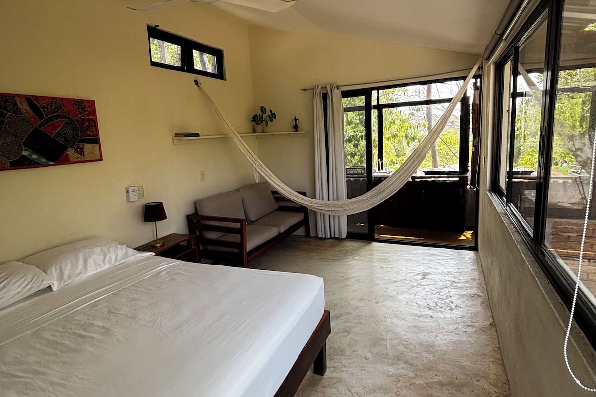 Une chambre avec un lit, un hamac, un ventilateur de plafond, une fenêtre et une vue sur la verdure.