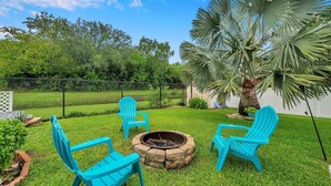 Un patio trasero con un brasero, tres sillas Adirondack azules y una palmera.