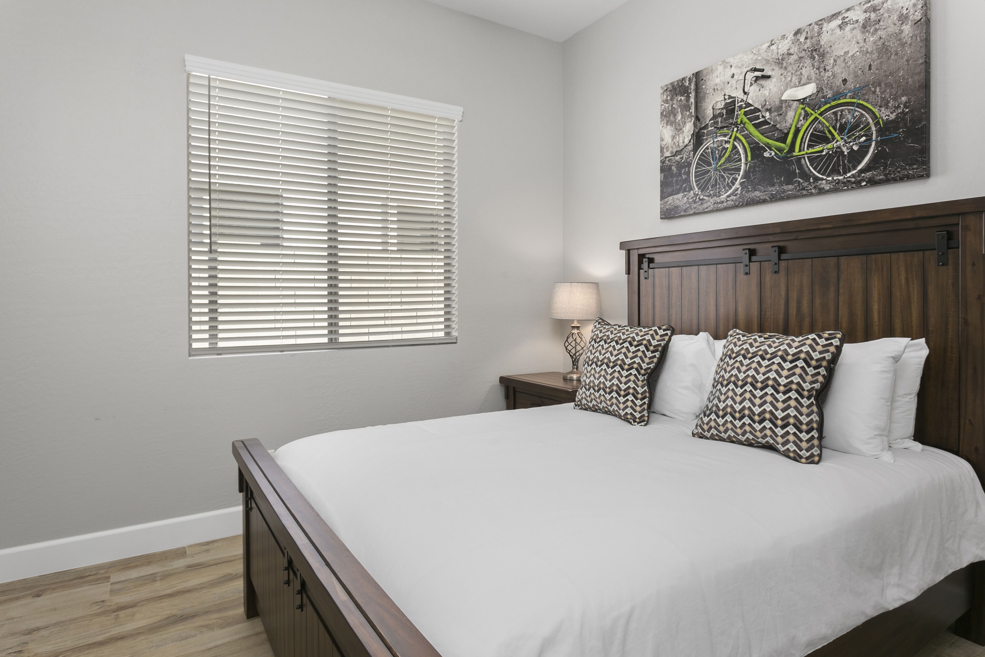 A bedroom with a bed, a nightstand, a lamp, a window with blinds, and a wall-mounted bicycle painting.