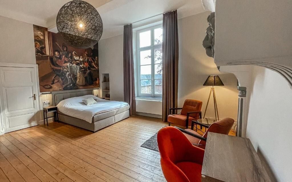 A modern hotel room with a large bed, a red armchair, a floor lamp, and a painting on the wall.