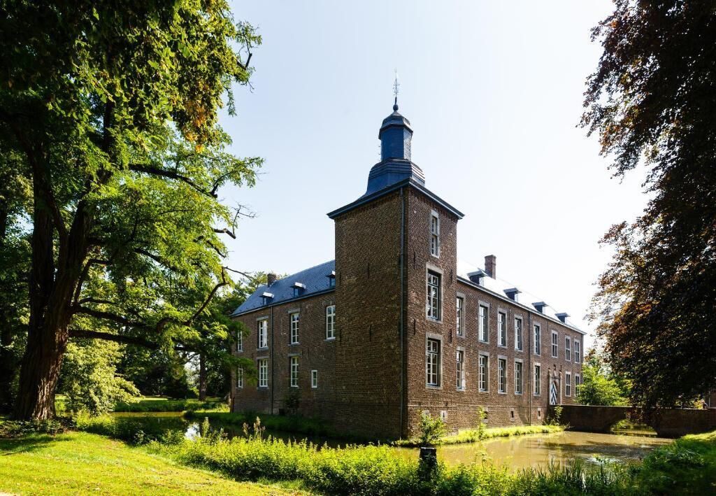 A historic brick building with a prominent tower, surrounded by lush greenery and a calm water body.