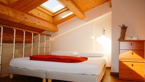 Un dormitorio con tragaluz, vigas de madera en el techo, una cama con ropa blanca y una manta roja, un aparador de madera y una pared blanca.