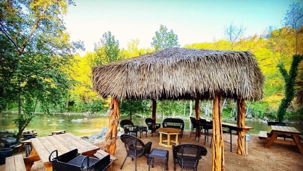 Un espacio para comer al aire libre con techo de paja, mesas de picnic y sillas, rodeado de árboles y un río.