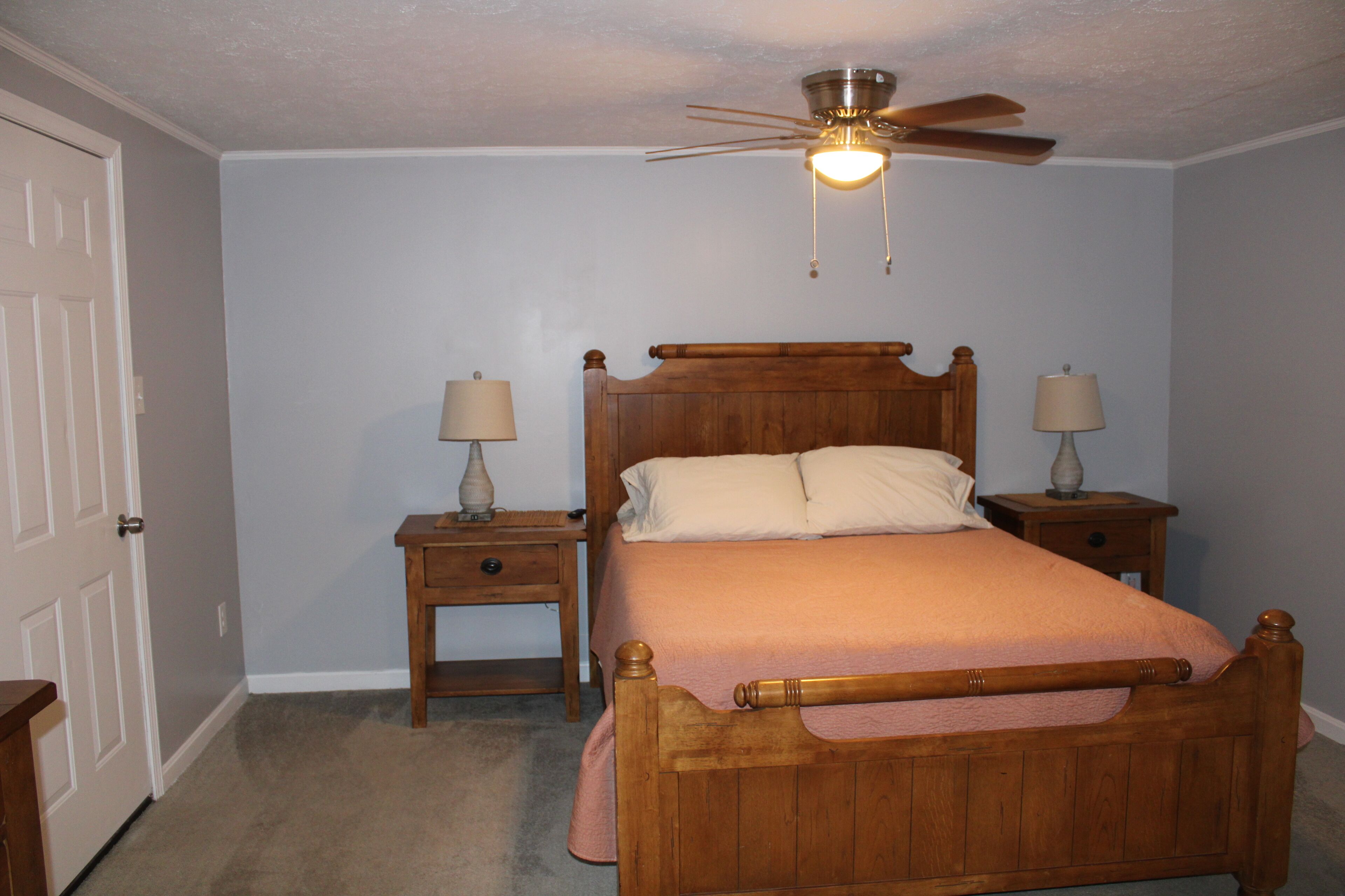 Une chambre à coucher avec un lit en bois, deux tables de chevet avec des lampes, un ventilateur de plafond et une porte.