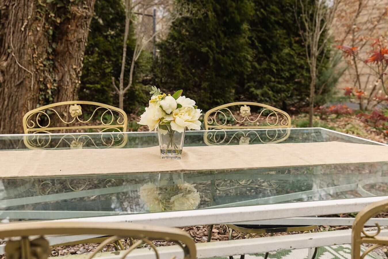 A glass-top table with two chairs, a vase with flowers, and a tree in the background.