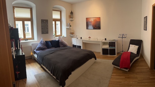 Un dormitorio con cama, escritorio, silla, radiador, ventana y estante fijado a la pared.