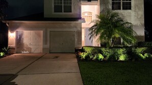 A two-story house with a garage, a well-lit front entrance, and a landscaped front yard with a palm tree. - LUXURY WINDSOR HILLS 4 BR 5BA POOL & SPA HOME 2.5 MILES to DSNY +++ (Kissimmee)