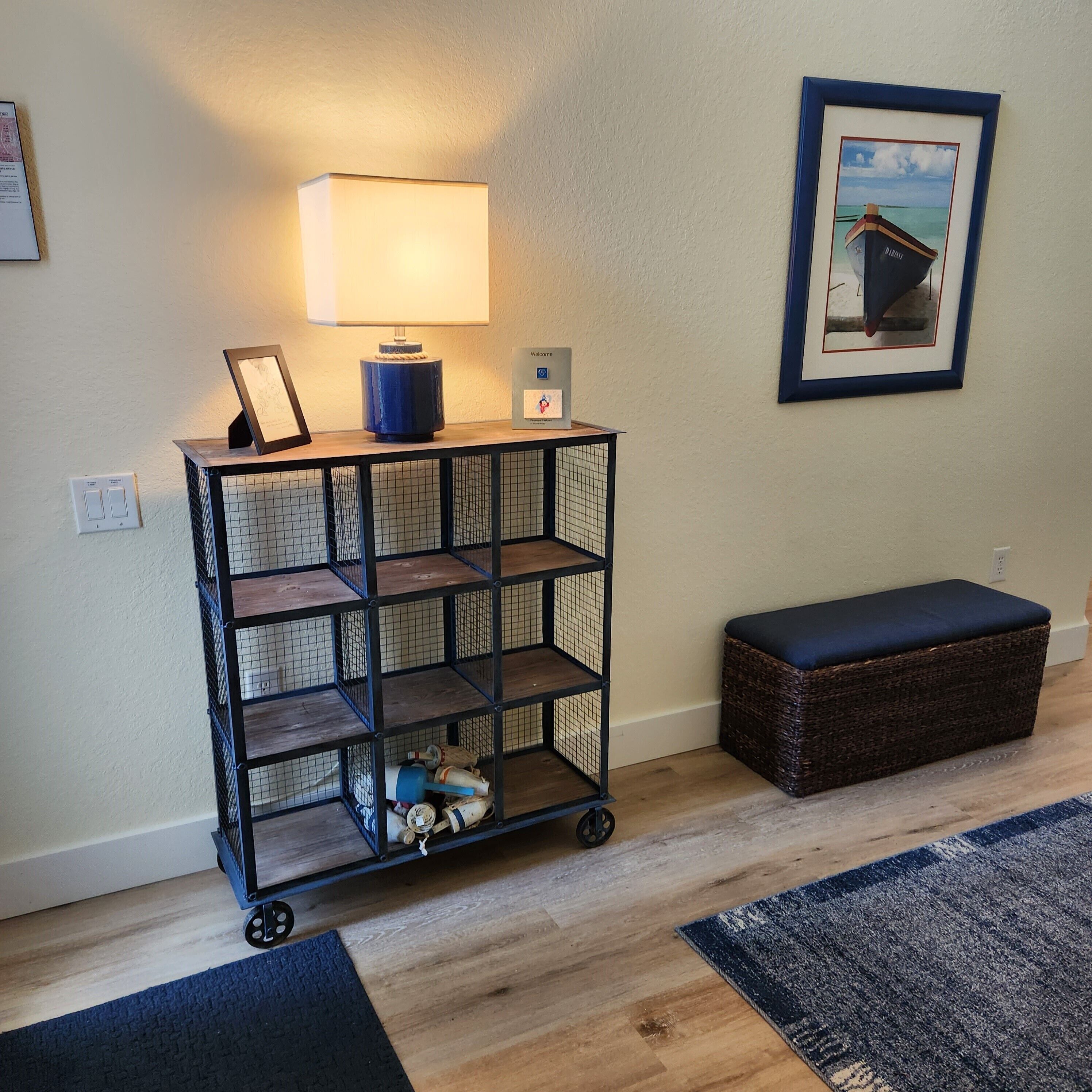 A room with a wooden floor, a black metal cart with shelves, a blue cushion bench, a framed picture on the wall, and a lamp on a table.