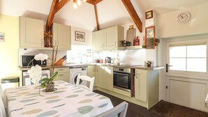 A kitchen with wooden beams, a dining table with a leaf-patterned tablecloth, a washing machine, and a clock on the wall. - Larch Barn (Helston)