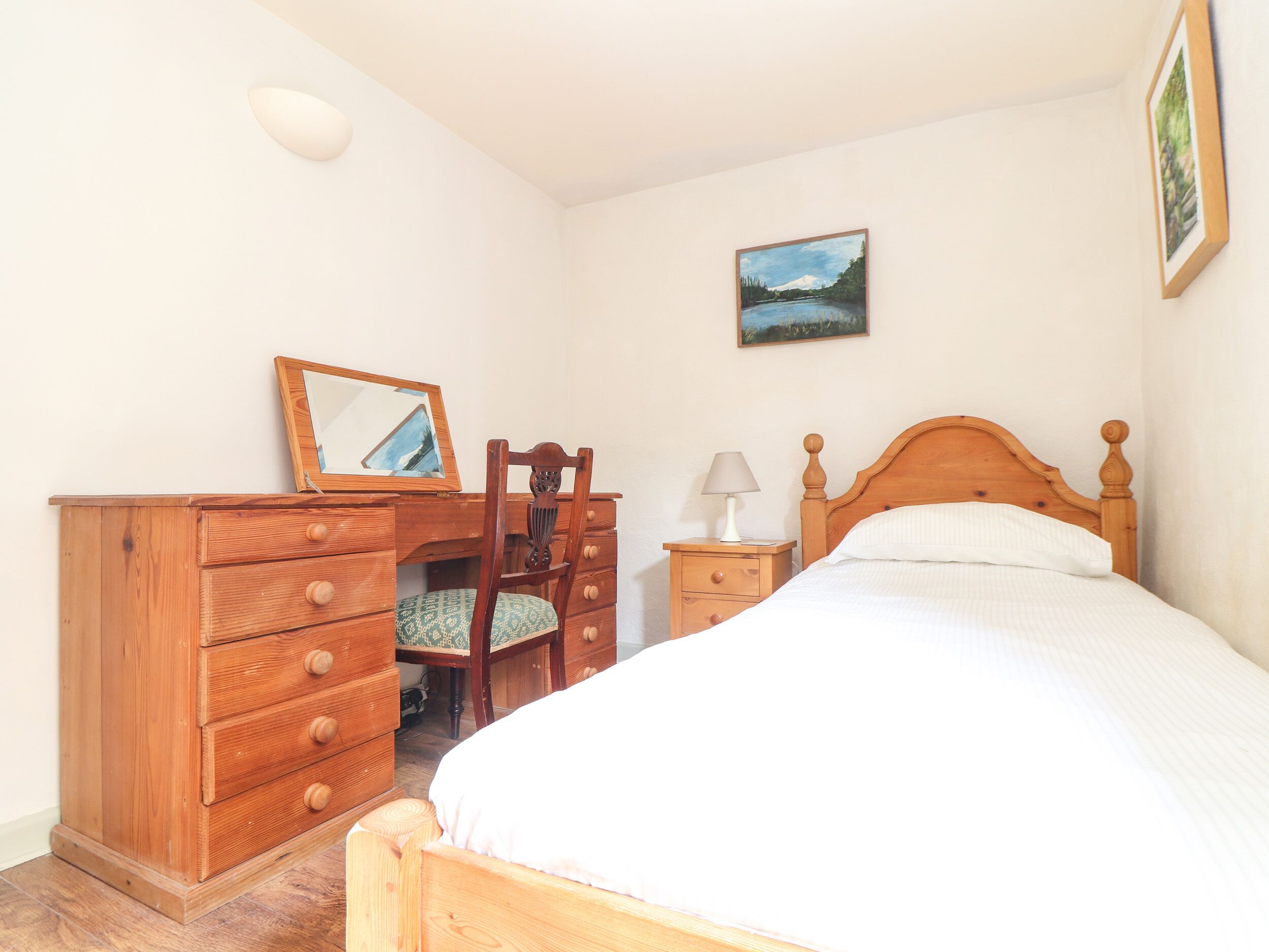 A bedroom with a wooden bed, a dresser with drawers, a chair, a nightstand with a lamp, and a painting on the wall.