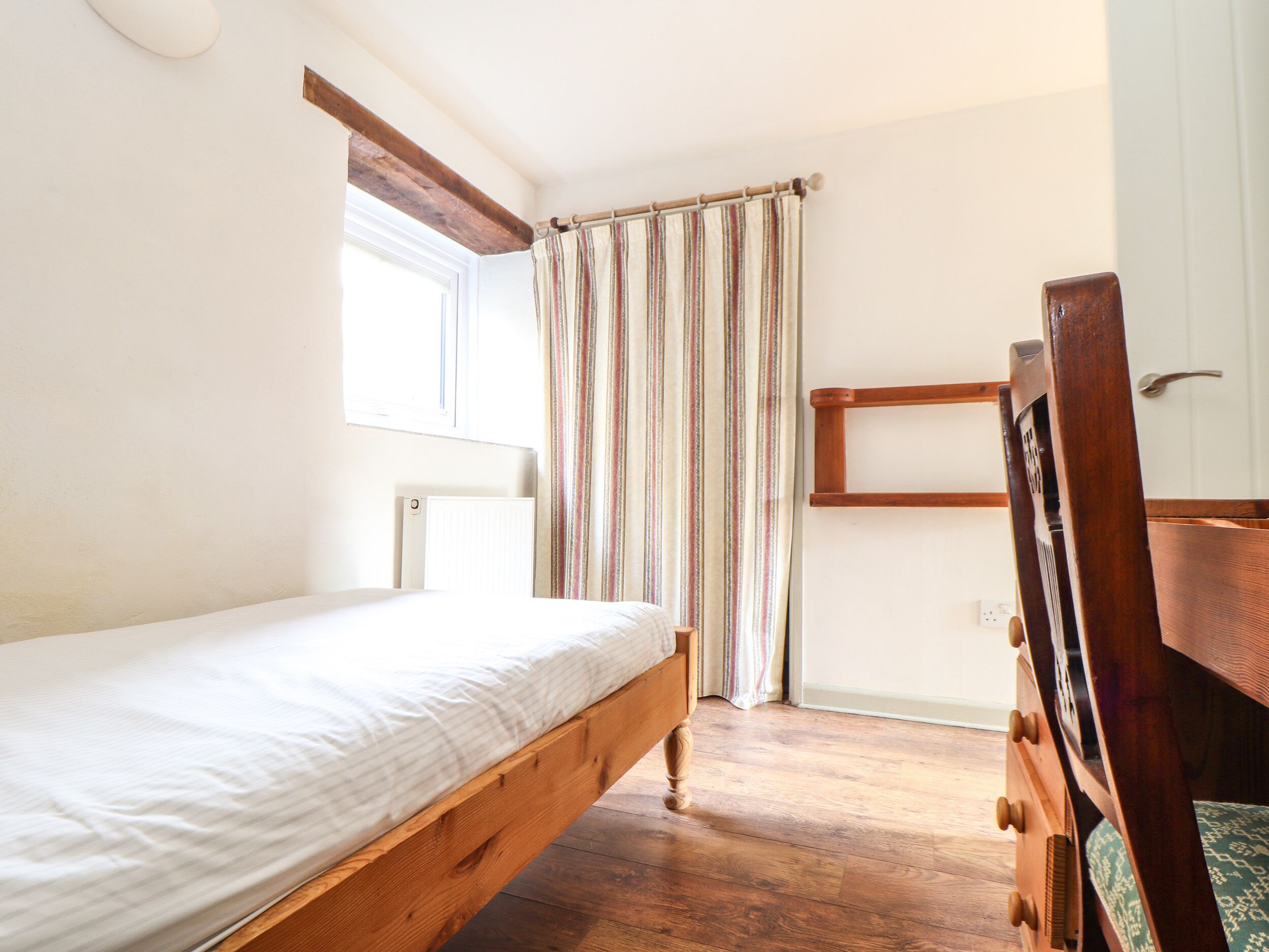 A bedroom with a wooden bed, a window, a radiator, a curtain, and a wooden chair.
