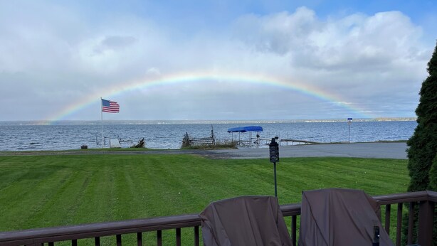 A deck with a table and chairs, overlooking a body of water with a rainbow and an American flag.