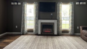 A living room with a fireplace, two windows with patterned curtains, a flat-screen TV mounted above the fireplace, and a brown leather sofa.