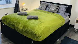A modern bedroom with a green bedspread, black headboard, and a view of a residential area through a window.